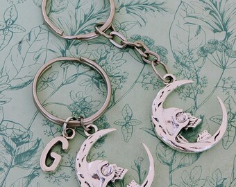 May include: Two silver keychains with a crescent moon design. Each moon has a skull inside and one keychain has a silver charm with the letter G.