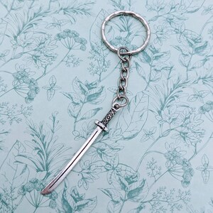 May include: Silver keychain featuring a small, detailed sword charm. The sword has a silver blade and a black handle with intricate designs.