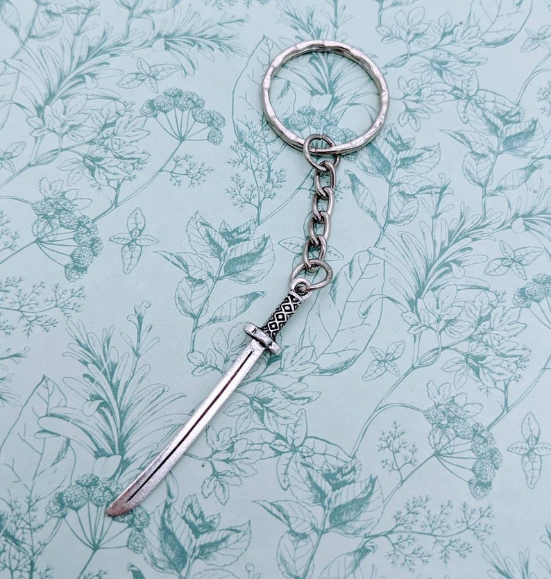 May include: Silver keychain featuring a small, detailed sword charm. The sword has a silver blade and a black handle with intricate designs.