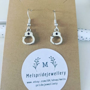 May include: Silver dangle earrings shaped like handcuffs. The earrings are on a brown card with the text "Mels pridejewellery www.etsy.com/UK/shop/mels pridejewellery"