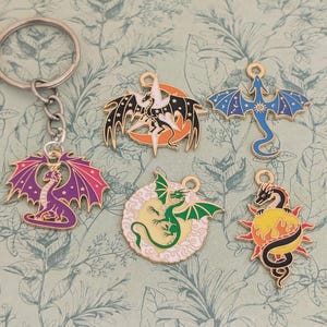 Könnte beinhalten: Eine Sammlung farbenfroher Drachenanhänger, darunter ein Schlüsselanhänger mit einem lila und rosa Drachen. Weitere Anhänger zeigen Drachen in Schwarz, Grün und Blau mit goldenen Akzenten. Der Hintergrund ist gemustert.