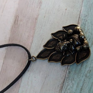 May include: A black cord necklace with a bronze pendant featuring a detailed depiction of the Hindu deity Ganesha sitting on a lotus flower.