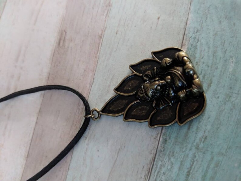 May include: A black cord necklace with a bronze pendant featuring a detailed depiction of the Hindu deity Ganesha sitting on a lotus flower.