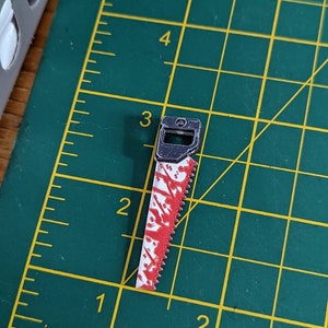 May include: A small, silver-colored saw with a red and white handle. The saw blade is covered in red paint splatters, giving it a bloody appearance.