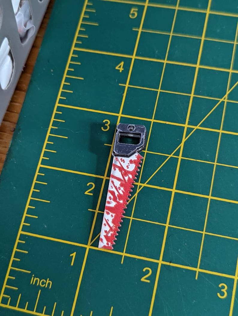 May include: A small, silver-colored saw with a red and white handle. The saw blade is covered in red paint splatters, giving it a bloody appearance.