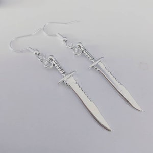 May include: A pair of silver dangle earrings featuring a sword design. The swords have a serrated edge and a pointed tip.