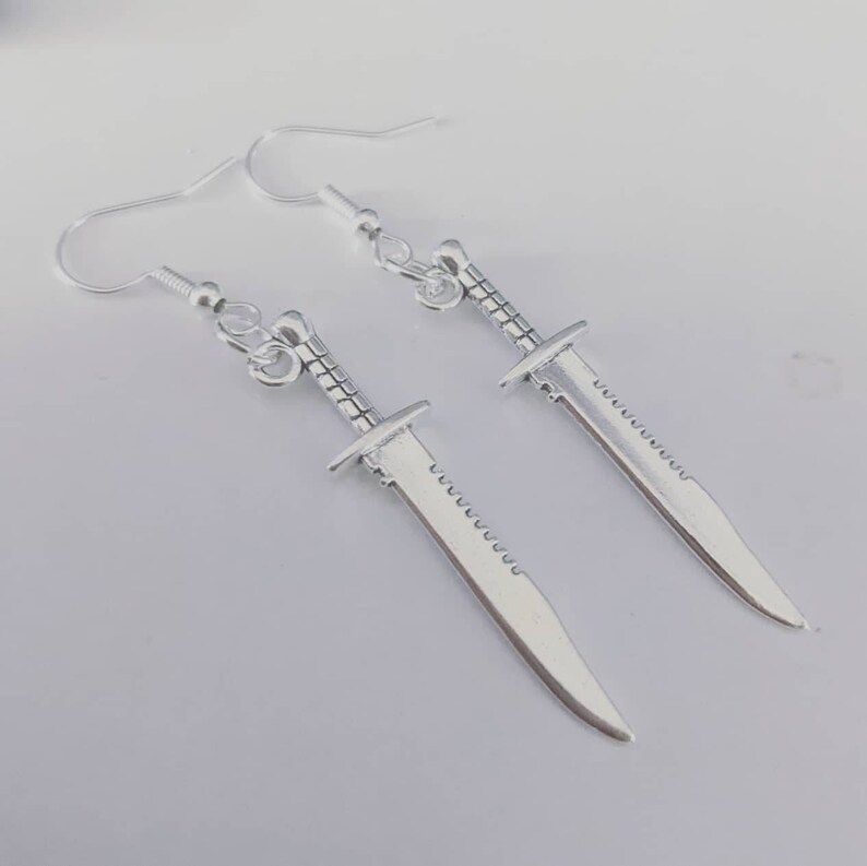 May include: A pair of silver dangle earrings featuring a sword design. The swords have a serrated edge and a pointed tip.