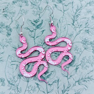 May include: A pair of pink snake earrings with white celestial designs. The earrings are made of acrylic and have silver hooks.