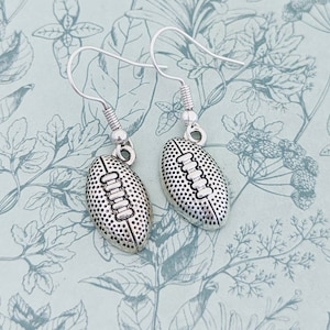 May include: Silver dangle earrings featuring a football design. The football is textured and has laces.