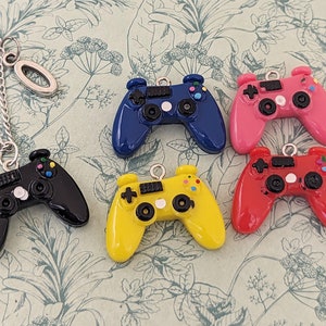 以下が含まれることがあります： 6つのカラフルなプラスチック製ゲームコントローラーチャーム。それぞれ異なる色（黒、青、黄、赤、ピンク、白）をしています。チャームは銀色のチェーンにぶら下がっています。