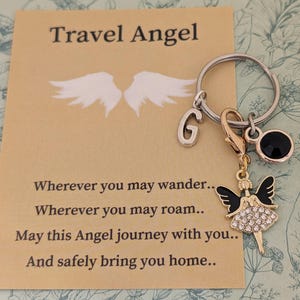 Op de afbeelding: Een sleutelhanger met een reisthema. De zilveren sleutelring heeft een bedel van een kleine engel met zwarte vleugels en een strass rok. Het bevat ook een zwarte edelsteen en de letter "G". Een kaart met de tekst "Travel Angel" staat op de achtergrond.