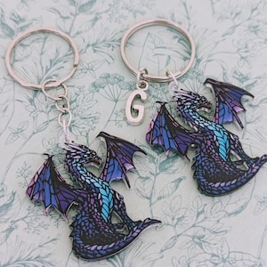 K&ouml;nnte beinhalten: Zwei Drachen-Schl&uuml;sselanh&auml;nger mit silbernen Ringen. Die Drachen haben lila, blaue und t&uuml;rkisfarbene Fl&uuml;gel und K&ouml;rper. Ein Schl&uuml;sselanh&auml;nger hat einen silbernen Buchstaben "G"-Anh&auml;nger.