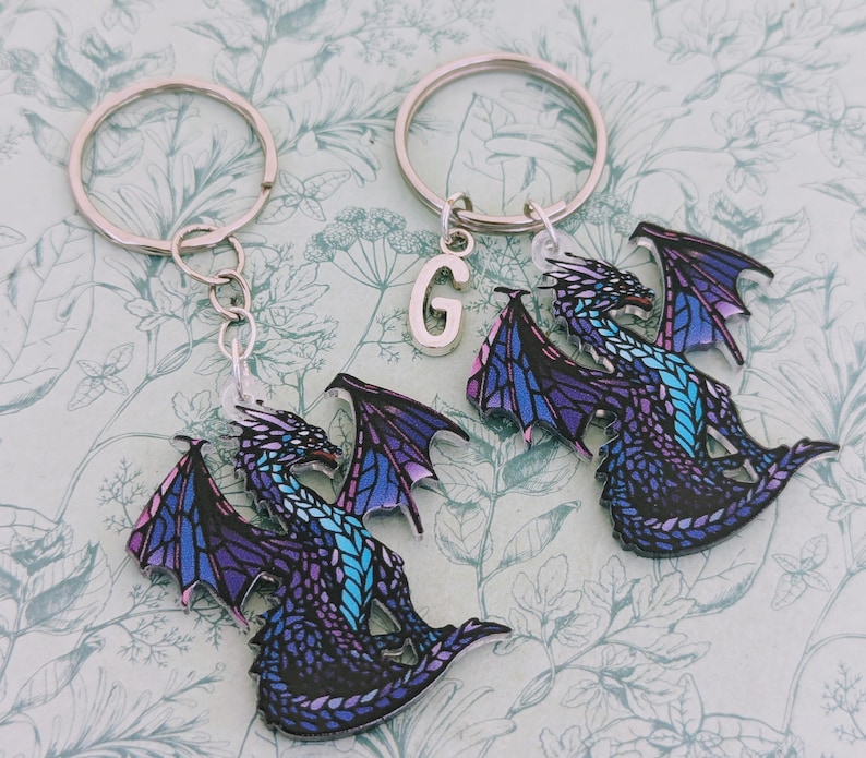 K&ouml;nnte beinhalten: Zwei Drachen-Schl&uuml;sselanh&auml;nger mit silbernen Ringen. Die Drachen haben lila, blaue und t&uuml;rkisfarbene Fl&uuml;gel und K&ouml;rper. Ein Schl&uuml;sselanh&auml;nger hat einen silbernen Buchstaben "G"-Anh&auml;nger.