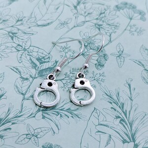 May include: A pair of silver dangle earrings featuring a handcuff design. The earrings are on a floral patterned background.