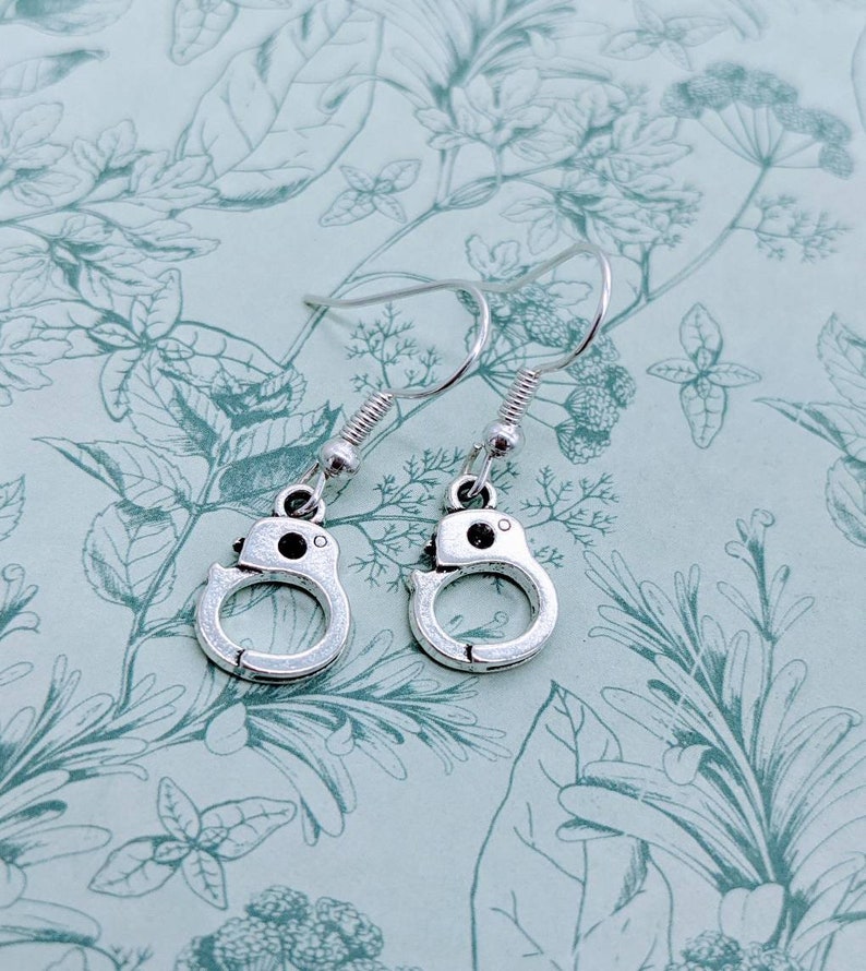 May include: A pair of silver dangle earrings featuring a handcuff design. The earrings are on a floral patterned background.