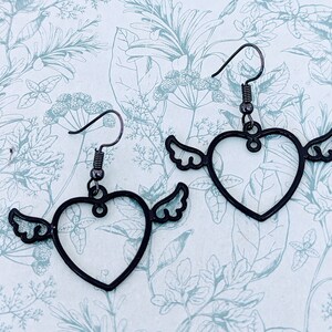 Könnte beinhalten: Ein Paar schwarze herzförmige Ohrringe mit Flügeln. Die Ohrringe sind aus Metall und haben ein schlichtes, elegantes Design.