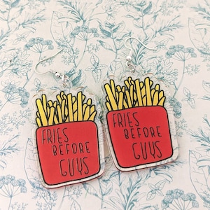 Puede incluir: Un par de pendientes de acrílico transparente con un diseño rojo y amarillo. Los pendientes presentan una imagen de dibujos animados de papas fritas en un recipiente rojo con el texto "Fries Before Guys".