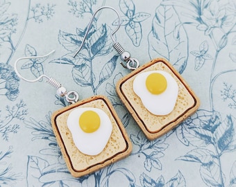 May include: A pair of novelty earrings featuring miniature toast slices topped with fried eggs. The earrings have a light brown toast base, a white egg white, and a yellow yolk. The earrings hang from silver-colored hooks.