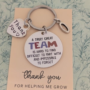 May include: A silver keyring with a heart-shaped charm that says "Thank You" and a round charm that says "A Truly Great TEAM is hard to find, difficult to part with, and impossible to forget." The keyring is on a card that says "Thank You for helping me grow."