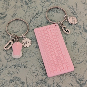 May include: Two keyrings with silver hoops. One features a pink computer mouse charm and a "BFF" charm. The other has a pink keyboard charm, a "G" charm, and a "BFF" charm. The background has a floral pattern.