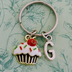Pode incluir: Um porta-chaves prateado com um berloque de cupcake e um berloque com a letra "G". O berloque de cupcake tem cobertura branca com granulado vermelho e verde, uma cereja vermelha e uma base dourada. O fundo é um padrão floral.