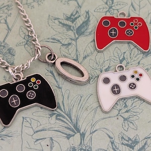 以下が含まれることがあります： 黒いコントローラーペンダントが付いたシルバーチェーンネックレスと、赤、白、黒の3つのコントローラーチャーム。 コントローラーには色の付いたボタンとプラス記号が付いています。 背景には花柄があります。