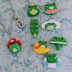 Könnte beinhalten: Eine silberne Halskette mit einem Pilzanhänger und mehreren grünen Frosch-Anhängern. Zu den Frosch-Anhängern gehören ein Frosch auf einer Seerose, ein Frosch auf einer Ente und ein Frosch in einem Schwimmring. Die Anhänger sind grün, rot, weiß und gelb.