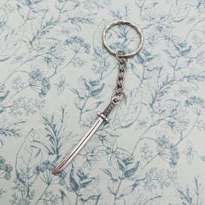 May include: A silver keychain with a sword charm. The sword has a detailed handle and a sharp point. The keychain is on a white background with a floral pattern.