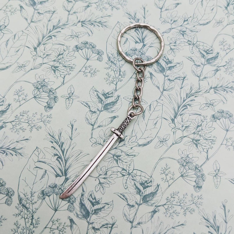 May include: A silver keychain with a sword charm. The sword has a detailed handle and a sharp point. The keychain is on a white background with a floral pattern.