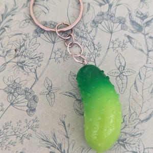 May include: A novelty keyring with a green and yellow pickle charm. The pickle has a textured surface and is attached to a silver chain and a rose gold key ring. The background has a floral pattern.