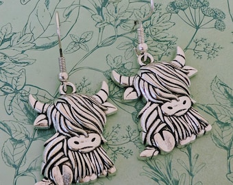May include: A pair of silver-colored Highland cow earrings. Each earring features a detailed, stylized cow design with horns, a shaggy mane, and a smiling face. The earrings hang from a simple hook.