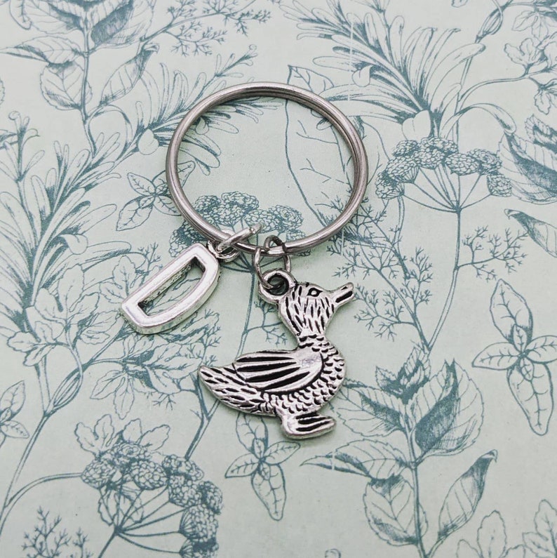 Op de afbeelding: Zilveren sleutelhanger met een eendenhanger en een letter D-hanger. De sleutelhanger bevindt zich op een groene en witte bloemenachtergrond.