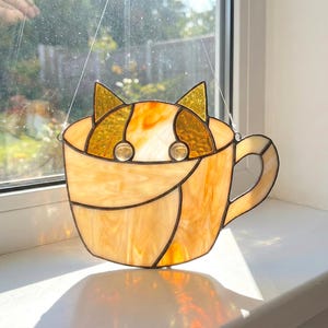 May include: Stained glass cat in a teacup suncatcher. The cat's ears and eyes are visible above the cup's rim. The teacup and cat are shades of amber and gold, with black lines outlining the design. The suncatcher hangs from a thin wire.