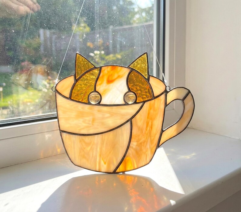 May include: Stained glass cat in a teacup suncatcher. The cat's ears and eyes are visible above the cup's rim. The teacup and cat are shades of amber and gold, with black lines outlining the design. The suncatcher hangs from a thin wire.