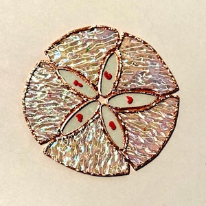 May include: A stained glass sand dollar with iridescent glass segments and copper foil edging. The center features six white petals, each with a red dot. The design is circular and reminiscent of a sea creature.