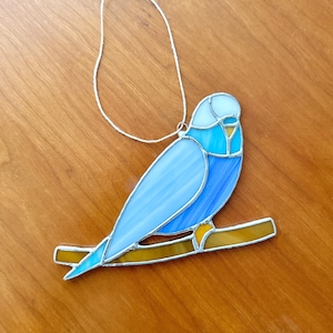 Op de afbeelding: Gebrandschilderde glazen parkiet suncatcher in blauwtinten, zittend op een gele tak. De vogel heeft een zilveren omtrek en een touwtje om op te hangen. Een decoratief stuk voor een raam of muur.