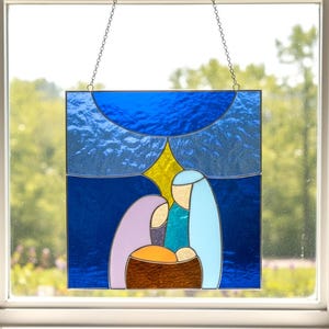 May include: Stained glass panel showing the Nativity scene. The artwork features a blue sky with a yellow star, and figures in purple, blue, and teal. The figures are positioned above a brown manger. The panel is framed in a dark blue border and hangs from a chain.