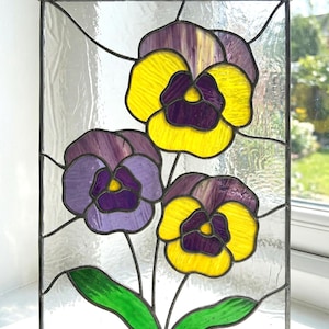 May include: Stained glass panel featuring three pansy flowers in shades of yellow, purple, and green. The flowers have a detailed design with a dark border. The panel is rectangular and hangs from a thin wire.