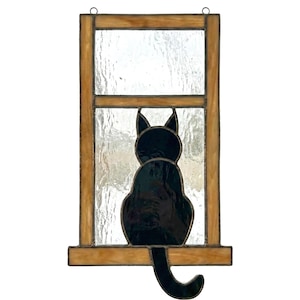 May include: Stained glass window art featuring a black cat silhouette. The cat is sitting, facing away, with its tail curled. The window frame is a warm brown color, and the glass has a textured appearance.