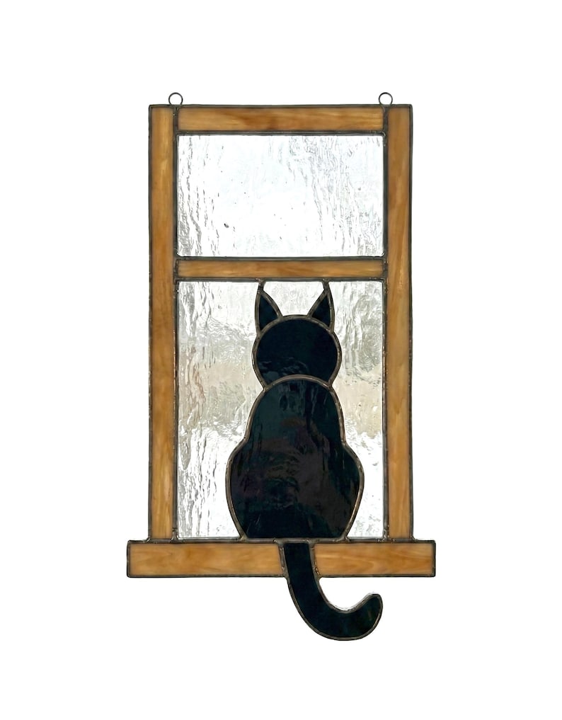 May include: Stained glass window art featuring a black cat silhouette. The cat is sitting, facing away, with its tail curled. The window frame is a warm brown color, and the glass has a textured appearance.