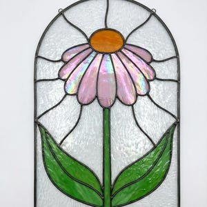 May include: Stained glass panel featuring a stylized flower with pink iridescent petals and an orange center. The flower has a green stem and two green leaves. The panel is framed in black and has a textured, white background.