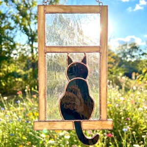 May include: Stained glass window panel featuring a black cat silhouette. The cat is perched on a ledge within a rectangular frame. The frame has a golden hue, and the glass is clear. The background is a sunny outdoor scene with flowers.