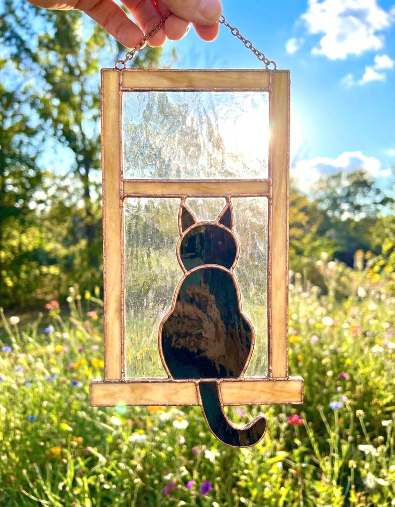 May include: Stained glass window panel featuring a black cat silhouette. The cat is perched on a ledge within a rectangular frame. The frame has a golden hue, and the glass is clear. The background is a sunny outdoor scene with flowers.