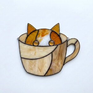 May include: Stained glass art piece featuring a cat peeking out of a coffee cup. The cat has orange and white fur, with amber-colored eyes. The cup is a light beige color with a brown handle and trim.