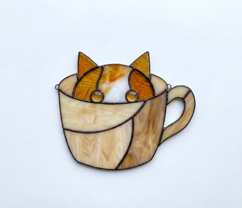 May include: Stained glass art piece featuring a cat peeking out of a coffee cup. The cat has orange and white fur, with amber-colored eyes. The cup is a light beige color with a brown handle and trim.