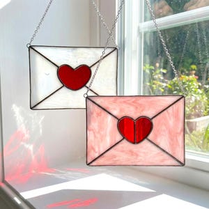 Peut inclure: Deux décorations en vitrail en forme d'enveloppe, une blanche et une rose, chacune avec un cœur rouge au centre. Les enveloppes sont soulignées de noir et suspendues par des chaînes argentées. L'enveloppe rose a un effet marbré.