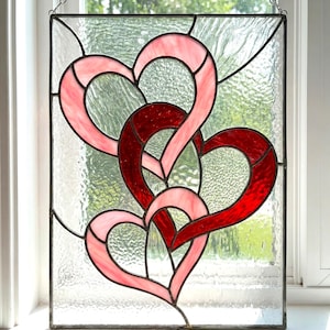 May include: Stained glass panel with three interlocking hearts in red and pink hues. The design is framed by black lines and set against a textured clear glass. A decorative piece for windows or walls, suitable for interior decor.