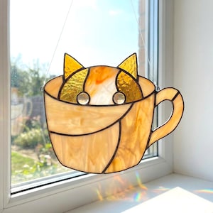 May include: Stained glass cat in a teacup suncatcher. The cat's ears and face peek over the rim of the cup. The teacup is a light amber color with a handle. The cat's eyes are two clear, round glass beads.