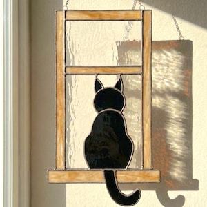 May include: Stained glass window art featuring a black cat silhouette sitting in a window frame. The frame has a light brown color, and the glass is clear. The cat is facing away from the viewer, with its tail curled.