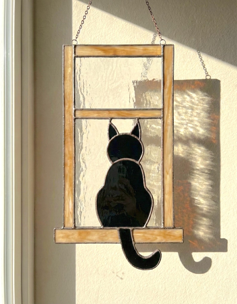 May include: Stained glass window art featuring a black cat silhouette sitting in a window frame. The frame has a light brown color, and the glass is clear. The cat is facing away from the viewer, with its tail curled.